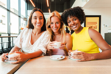 Group of young women drinking a coffee cup on a bar or restaurant. Three multiracial girls holding a mug looking at camera. Females smiling and laughing on a coffeeshop. Ladies resting with a drink