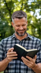 Obraz premium A young man stands in a park, holding an open Bible in one hand and a smartphone in the other, surrounded by trees and greenery on a sunny day