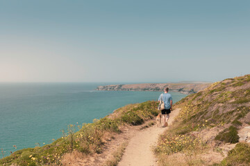 Caucasian casual attire man walking Whippet dog along clifftop coastal path towards Bedruthan Steps with stunning sea views and green hills on a sunny day, overlooking the sea Cornwall, UK