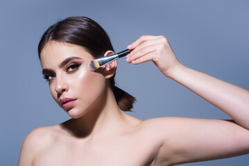 Beautiful young woman applying makeup on studio background. Pretty girl holding makeup brushes and make up on face with cosmetics. Facial beauty. Perfect skin, natural make up.