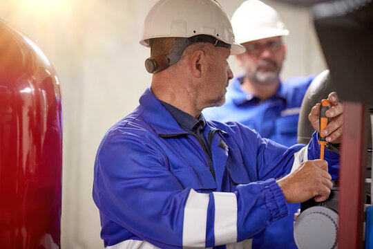 Two Engineers wearing blue overalls and white hard helmets performing  boiler room inspection and preventive maintenance. 