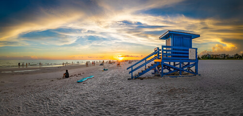 Panoramic photo of a sunset at a beach with a blue lifeguard tower, capturing the lively atmosphere and colorful sky as beach goers enjoy the evening. Perfect for travel, landscape, and decor use.