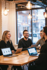 A group of people are sitting around a table with laptops open, smiling and laughing.