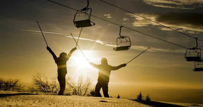 Silhouette of two friends against sunset sky raising their arms in celebration beneath a row of empty ski lift chairs. Skiers and chairlifts in winter resort. 50 fps on cinema camera