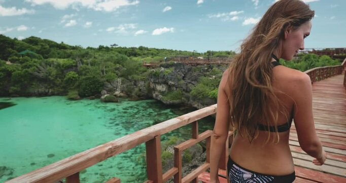 Woman enjoying a stunning view of a turquoise lagoon on a wooden bridge on a sunny day. Relaxing in nature, she embraces the tranquil vacation moment