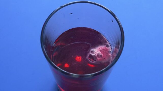 An effervescent tablet dissolves in a glass of water which turns burgundy, top view. Medium shot.
