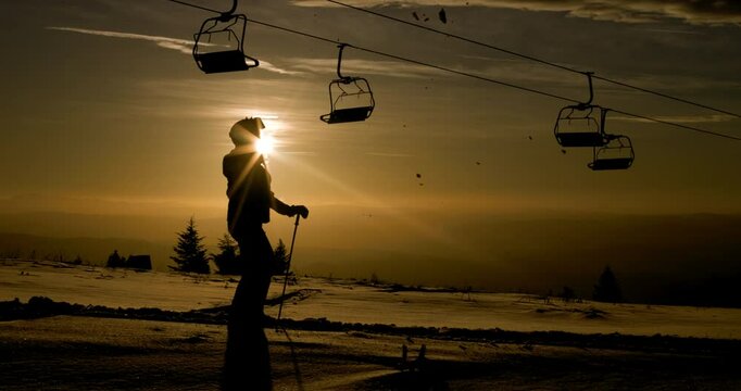 Silhouette of a female skier against sunset sky beneath a row of empty ski lift chairs. Girl throwing show in the air with her ski. 120fps cinema camera.