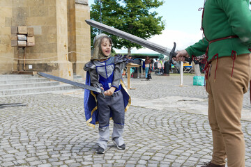 Boy in a knight costume with a sword, participating in a medieval reenactment outdoors. Enthusiastic and engaged in the activity, surrounded by other participants and historical elements