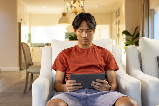 Sitting on armchair, young asian man using tablet in modern living room