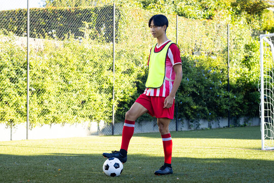 Young asian male Soccer player in red uniform standing on field with foot on ball