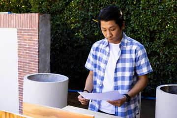 Reviewing blueprints, asian man in checkered shirt working on outdoor construction project