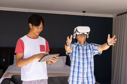 Using vr headset, asian father and teenage son holding tablet in bedroom