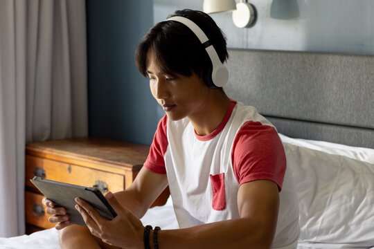 Using tablet and wearing headphones, asian man relaxing on bed in bedroom - Powered by Adobe