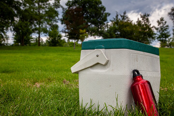 White cooler box with green lid and a red water bottle on a green lawn in a park with trees in the background