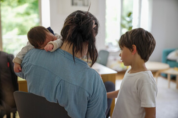 Fototapeta premium Mother holding sleeping baby on shoulder while talking to older child, indoor scene, family interaction, baby in white onesie, casual home environment, maternal care, bonding moment