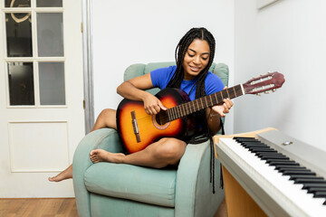 A 20-year-old dark-skinned girl with braids is playing a flamenco guitar on the couch at home. Concept of Latin women playing an instrument.