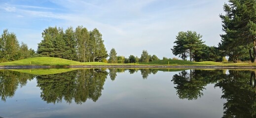 A golf green with a white flag near a pond, reflecting trees and the sky in the water. Peaceful scene with natural colors.