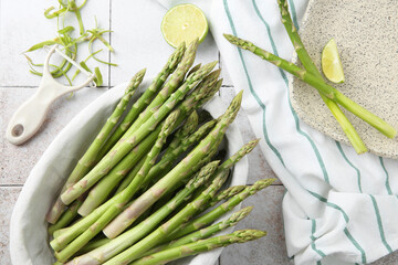 Fresh green asparagus stems, lime and vegetable peeler on light textured tiled table, flat lay