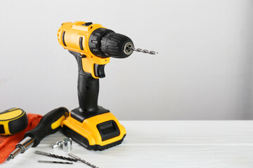 Cordless electric drill, gloves, bits, screwdriver and measuring tape on white wooden table, space...