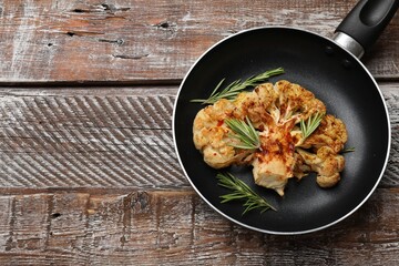 Tasty cauliflower steak and rosemary in frying pan on wooden table, top view. Space for text