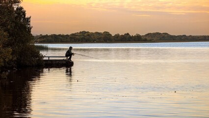 Silhouetted man fishing at sunset from pier at Ballyquirke lough, Galway, Ireland, sport and activities, catch and release, nature background