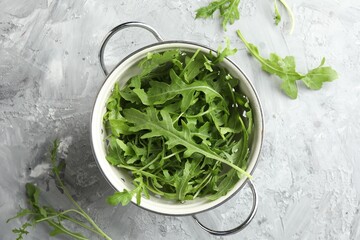 Fresh green arugula leaves in colander on grey textured table, top view