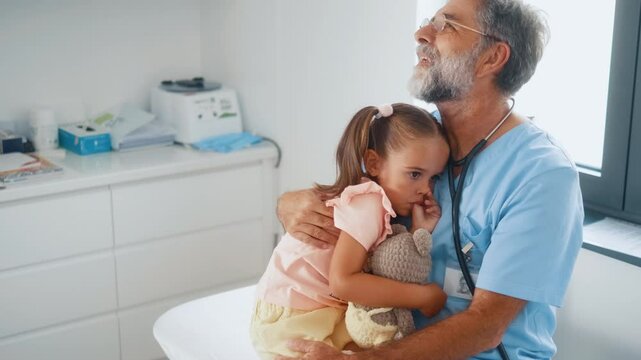 Elderly pediatrician hugs and caresses child patient little girl who is feeling insecure and scared at hospital examination