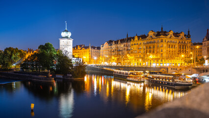 The Sitkov Water Tower stands tall along Masaryk Embankment in Prague, Czechia, as the sun sets and the city lights begin to illuminate the scene. The calm water reflects warm glow of the buildings.