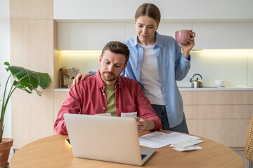 Serious man manages family budget frown looks to unpaid bills sits with laptop wife stands beside watch by husband. Focused married couple check account in banking app find mistake at credit payment.
