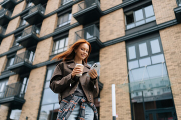 Low-angle view of stylish cheerful young woman standing on city street while drinking coffee and browsing smartphone with smile on face, posing on blurred background of modern building. © dikushin