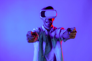 Gamer wear virtual reality glass while pointing two hands at camera. Happy caucasian man with VR goggles future innovation technology to enter virtual world simulation with neon background. Deviation.