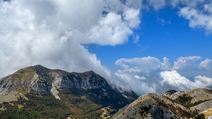  A scenic view of a mountain range with lush greenery and cloud-covered peaks, under a bright blue sky.