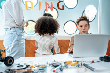 Kids in classroom learn engineering in the STEM class. Schoolboy sitting on chair and coding remote car with laptop.Teacher and other children are around him.Notebook, electric wire. Erudition.