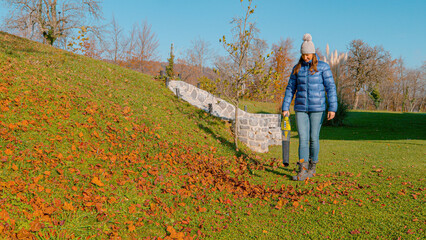Young woman using a portable leaf blower to clear dry fallen leaves from her maintained country...