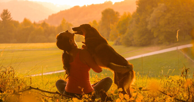 SILHOUETTE: Moment of bonding between lady and her playful pet as they sit by the golden lit meadow. Smiling woman and happy dog are enjoying the sunset after a walk through colorful autumn forest.