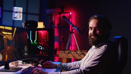 Portrait of smiling man in dark living room playing engaging video games on gaming PC at computer desk, chilling after work. Happy gamer contending against foes in online multiplayer shooter