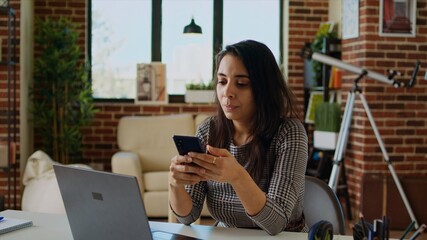 Smiling person checking mobile phone notifications at computer desk, taking break from work. Indian...