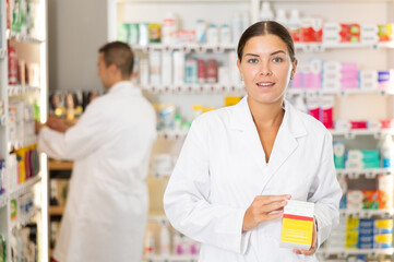 Kind young apothecary holding a box with remedy standing against shelves full of medicine in drugstore