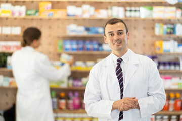 In pharmacy, positive male chemist stands and waits for visitors. Wide range of prescription and over-counter medications, patient care products, baby food