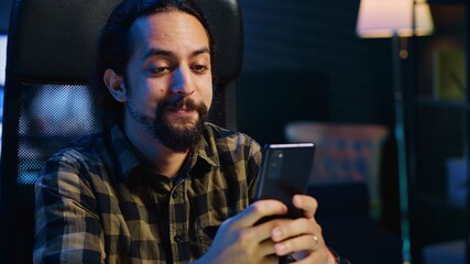 Smiling man at home texting best friend on smartphone, taking break while working in personal...