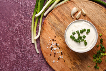 Board with bowl of tasty sour cream and sliced green onion on purple background