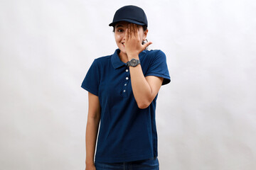Young asian woman courier wearing delivery uniform and cap covering one eye with hand, confident smile on face and surprise emotion.