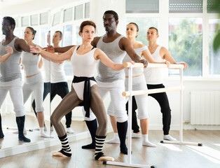 Group of multinational men and women practicing plie ballet movement at barre in dance studio