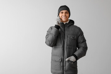Young man in puffer jacket talking by mobile phone on light background