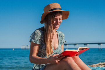 Only a young woman reading a book sitting on the sand of the beach. Blonde female enjoying the literature resting at shoreline. Caucasian girl relaxing with a novel in the seaside on her vacations