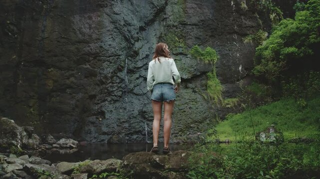 Tourist is standing on a rock, admiring a waterfall flowing on a volcanic rock wall. The lush vegetation surrounding the area creates a sense of tranquility and adventure