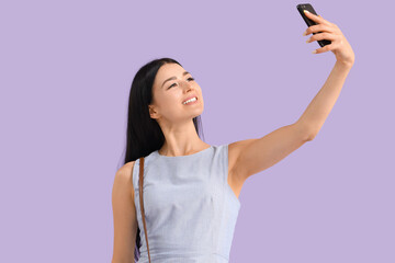 Young woman with mobile phone taking selfie on lilac background