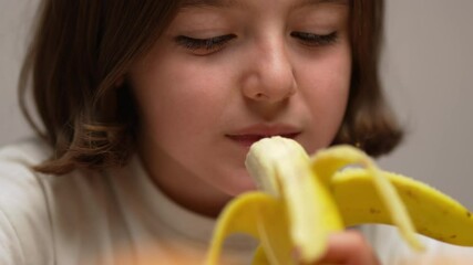 A close-up shot of a little girl taking a bite of a banana and eating it.​