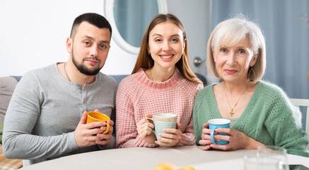 Cheerful elderly mother with adult son and his wife drinking tea, having pleasant talk and posing...
