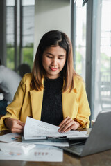 A young woman in a yellow blazer reviews documents at her desk in a modern office environment,...